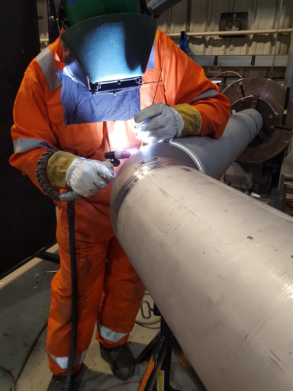En person i oransje vernedrakt og sveisehjelm sveiser et stort metallrør